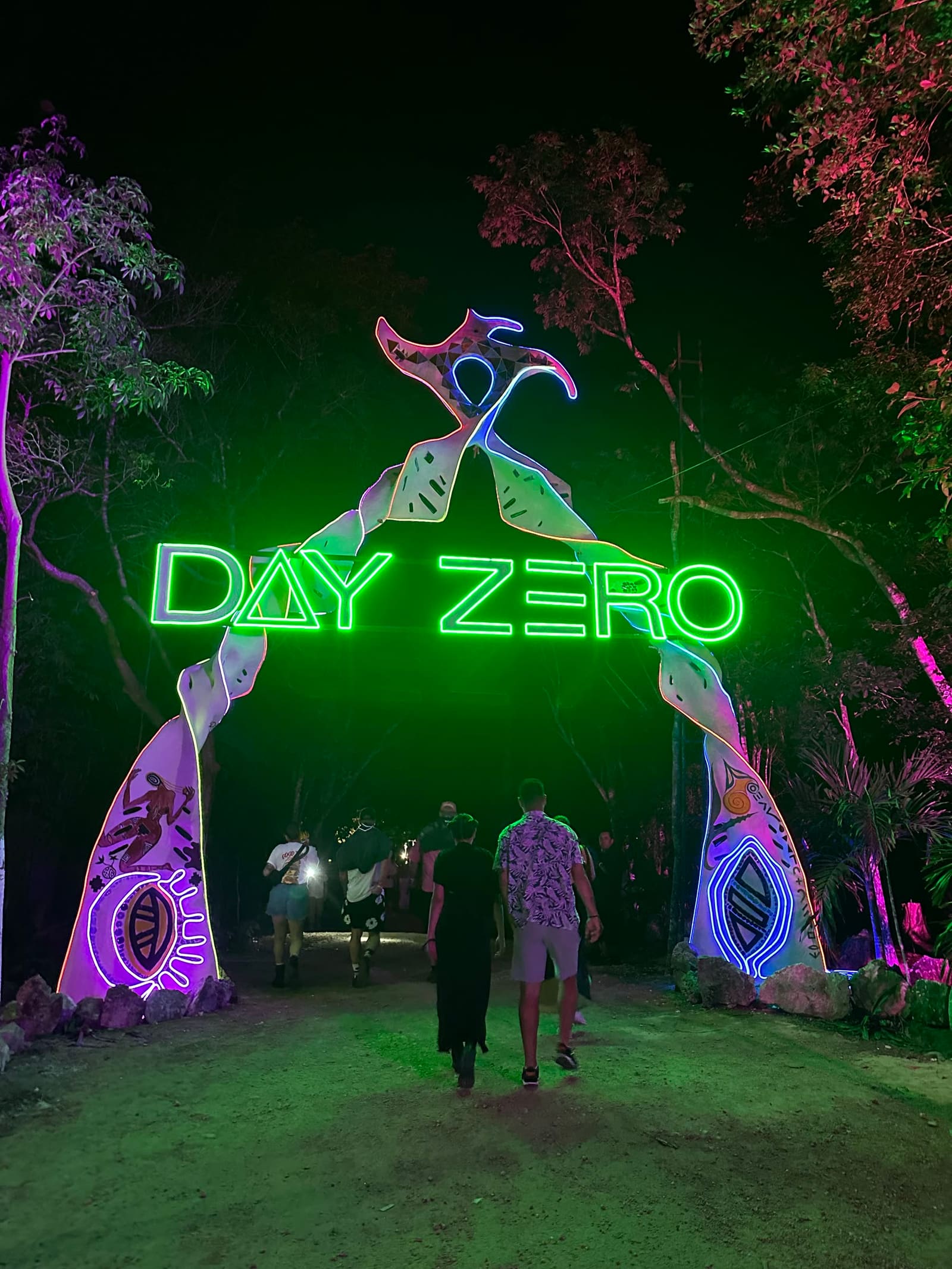 Day Zero entrance arch in the Tulum jungle, with green neon lettering and painted Maya figures, and people walking in at night