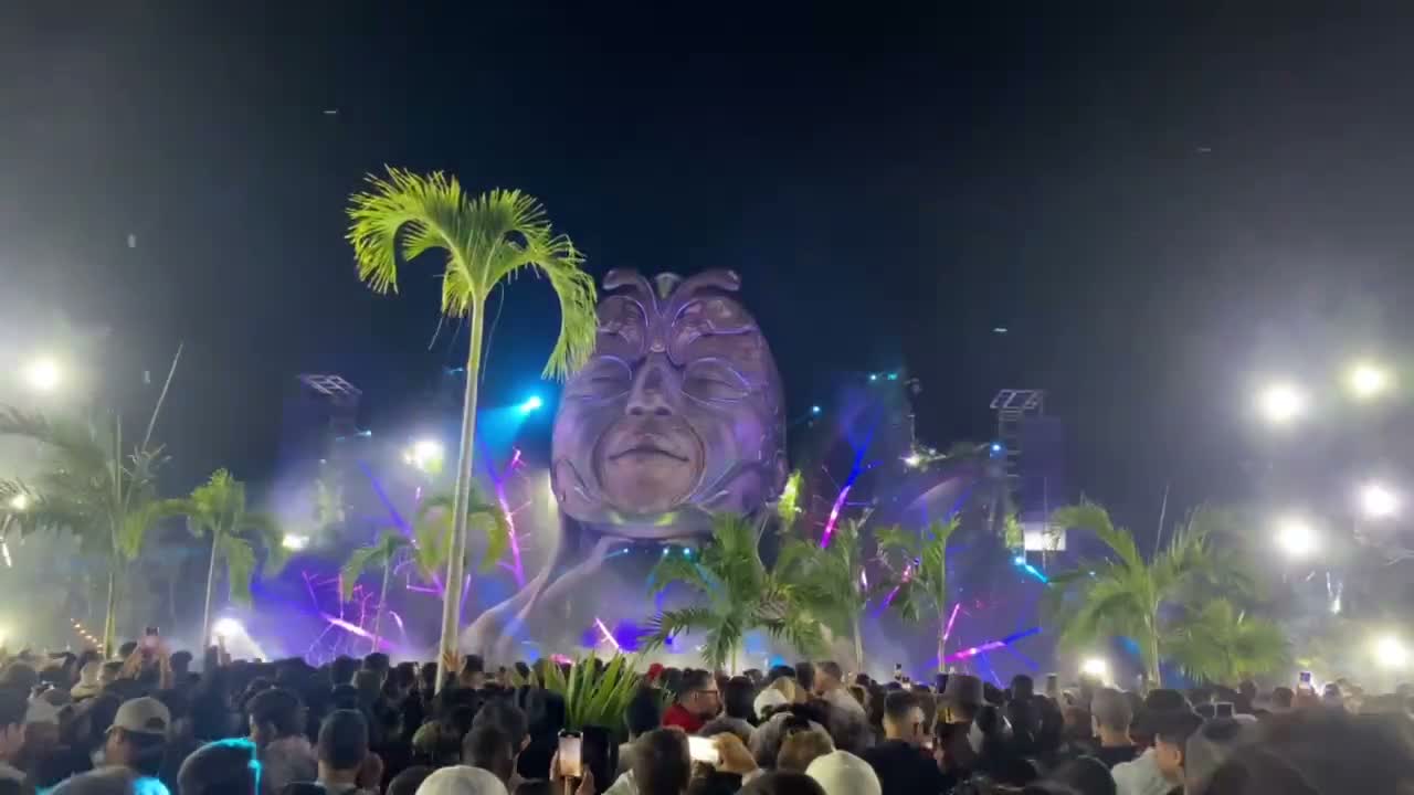 Sculpture of a giant head with closed eyes as the CORE stage in Tulum, among palm trees, lasers and a crowd at night